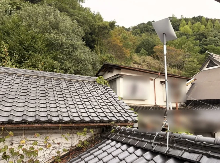 古民家の瓦屋根にポールで立てたスターリンクのアンテナと、裏山の林